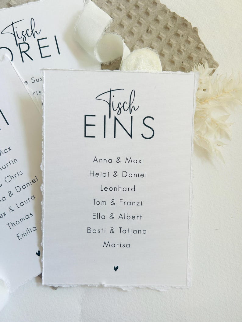K&ouml;nnte beinhalten: Eine wei&szlig;e rechteckige Karte mit B&uuml;ttenrand, die den schwarzen Aufdruck Tisch EINS sowie eine Namensliste mit Anna & Maxi, Heidi & Daniel, Leonhard, Tom & Franzi, Ella & Albert, Basti & Tatjana und Marisa und ein kleines Herz zeigt.