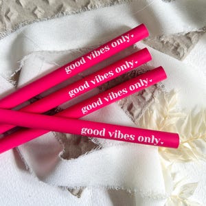 Könnte beinhalten: Vier leuchtend pinke Bleistifte mit dem weißen Schriftzug "good vibes only". Sie liegen diagonal auf einem strukturierten, neutralfarbenen Hintergrund mit weißen Stoffakzenten.