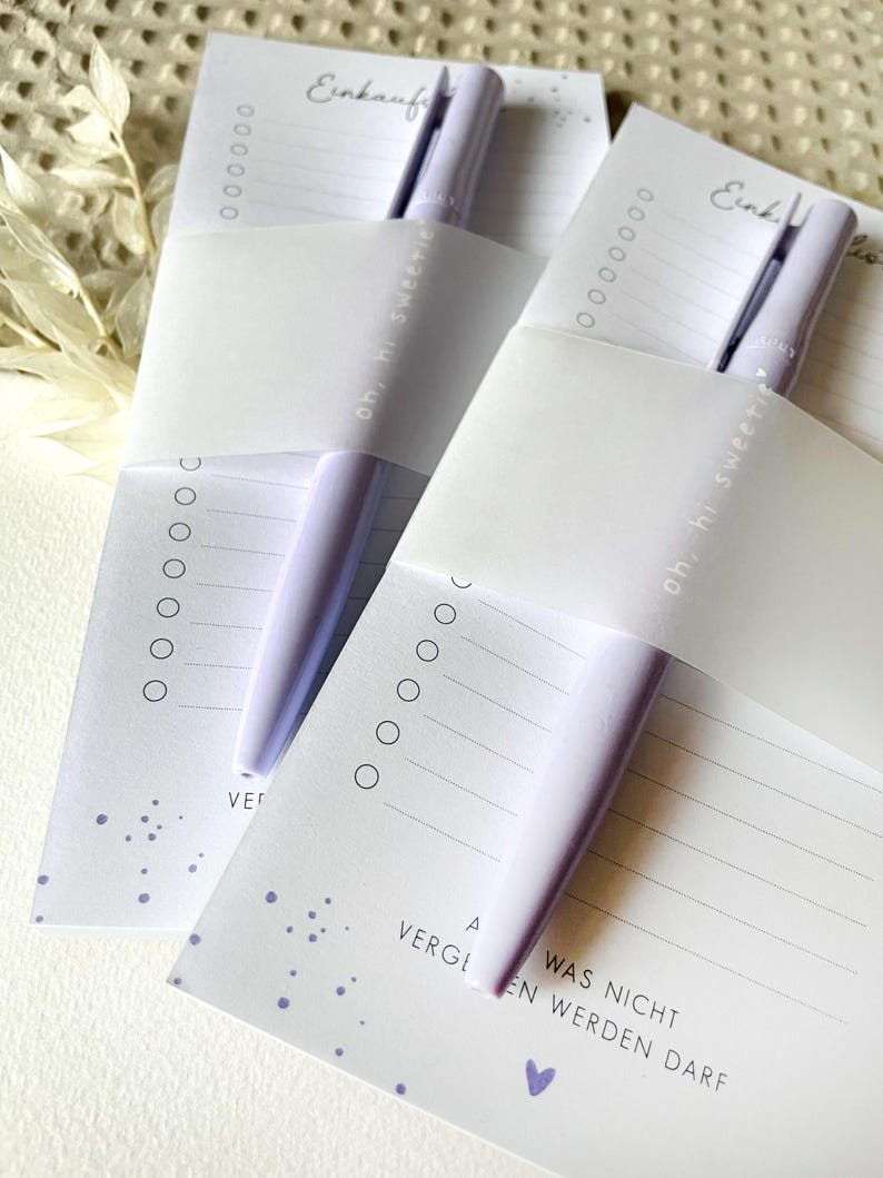 May include: Two white shopping list pads with purple pens. The pads have a dotted pattern and the text "Einkauf" and "oh, hi sweetie" on them. The text "WAS NICHT" and "VERGEN WERDEN DARF" is on the bottom of the pad.