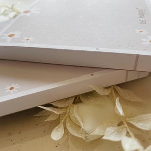 May include: Two stacks of white memo pads with a light gray background and a border of white daisies with brown centers. The top pad has the text "be happy" printed on the right side.