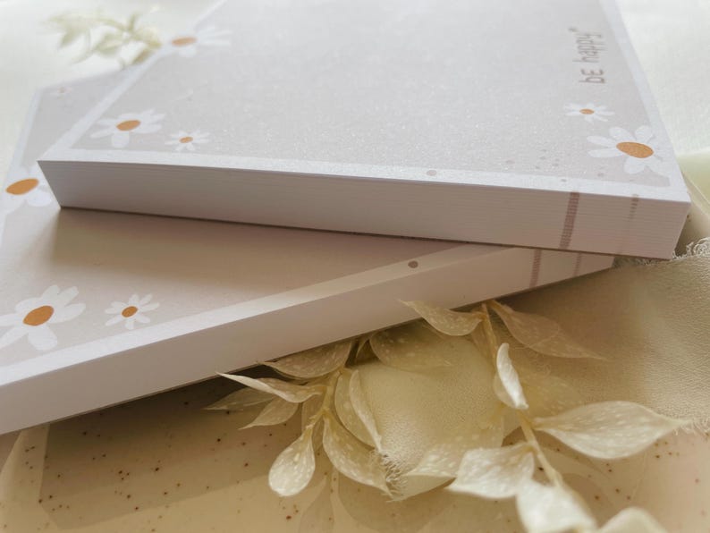 May include: Two stacks of white memo pads with a light gray background and a border of white daisies with brown centers. The top pad has the text "be happy" printed on the right side.