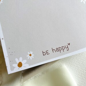 May include: A light gray card with a white border and a white daisy pattern. The card has the text "be happy" in brown lettering with a small heart symbol.