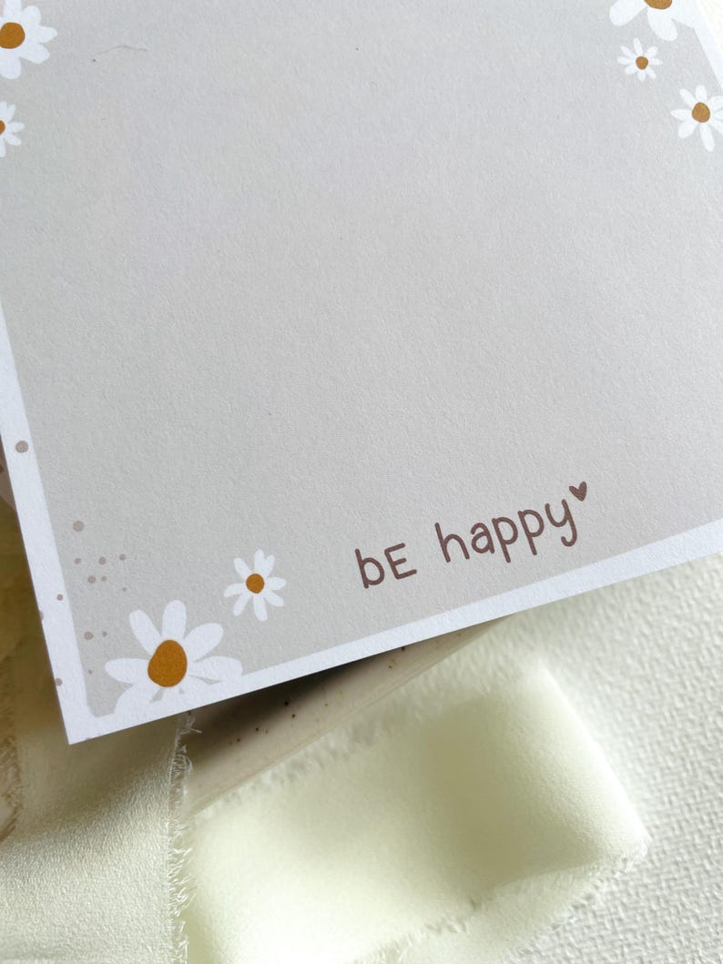 May include: A light gray card with a white border and a white daisy pattern. The card has the text "be happy" in brown lettering with a small heart symbol.