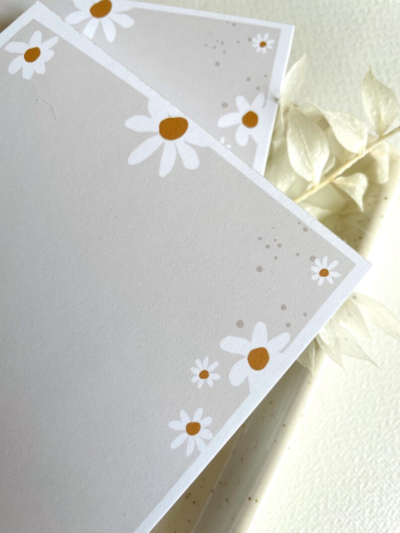 May include: Two gray cards with a white border and white daisies with yellow centers. The daisies are arranged around the edges of the cards.
