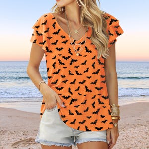 May include: Orange V-neck top with short, ruffled sleeves. The shirt is covered in a pattern of black bats. The model is wearing the shirt with denim shorts and gold jewelry. The background is a beach scene.