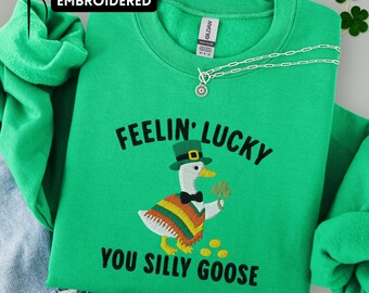 May include: Green sweatshirt with the embroidered text "FEELIN' LUCKY YOU SILLY GOOSE" and a cartoon goose wearing a hat and holding a shamrock. The word "EMBROIDERED" is above the sweatshirt.