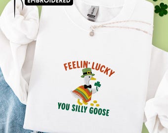 May include: White sweatshirt with an embroidered design featuring a goose wearing a green hat and poncho, holding a shamrock. The text reads "FEELIN' LUCKY YOU SILLY GOOSE". A gold necklace and the word "EMBROIDERED" are also visible.