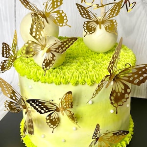 May include: A white cake decorated with green frosting and gold butterfly accents. The cake has a "Happy Birthday" topper and white chocolate balls with gold butterflies on top.
