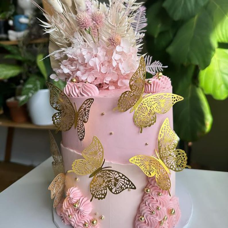 May include: A two-tiered pink cake decorated with gold butterfly accents and dried flowers. The cake is topped with a cluster of white hydrangeas.