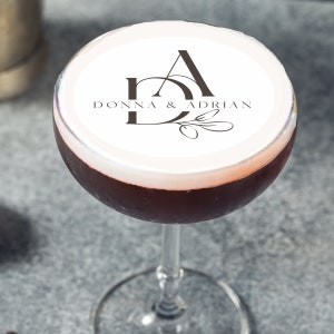 May include: A glass of red cocktail topped with white foam. The foam features a personalised design with the letters "D" and "A" intertwined and the text "Donna & Adrian" printed on top.
