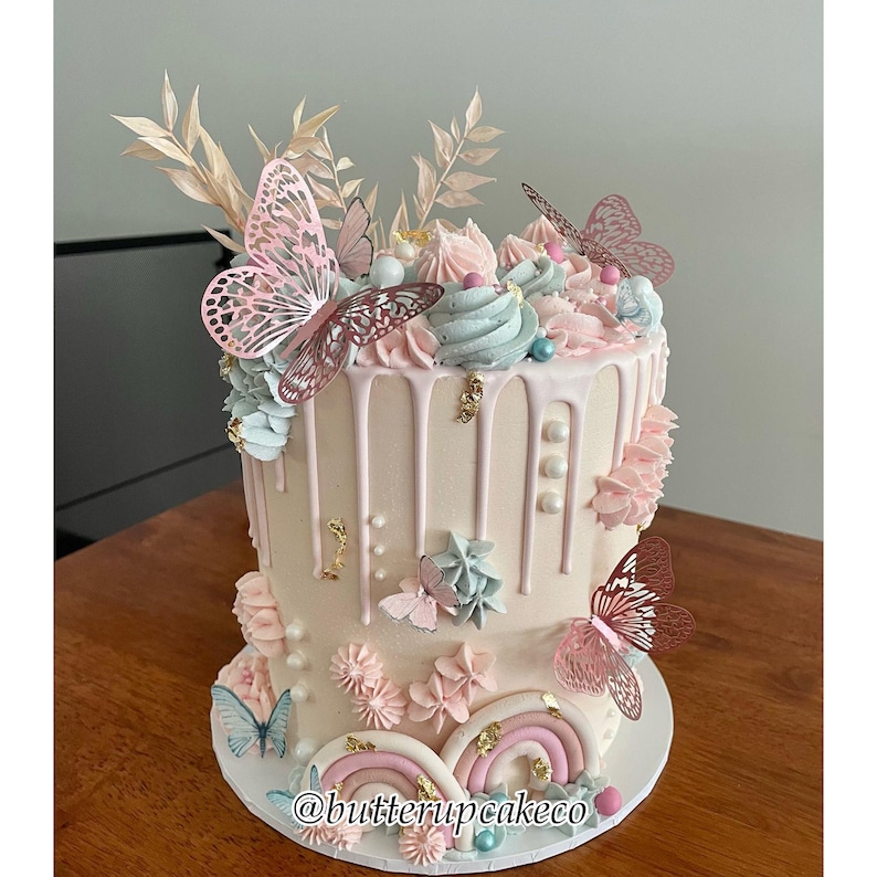 May include: A two-tiered cake decorated with pink and blue frosting, edible butterflies, and gold accents. The cake is on a white cake board with the text "@butterupcakeco" on the bottom.