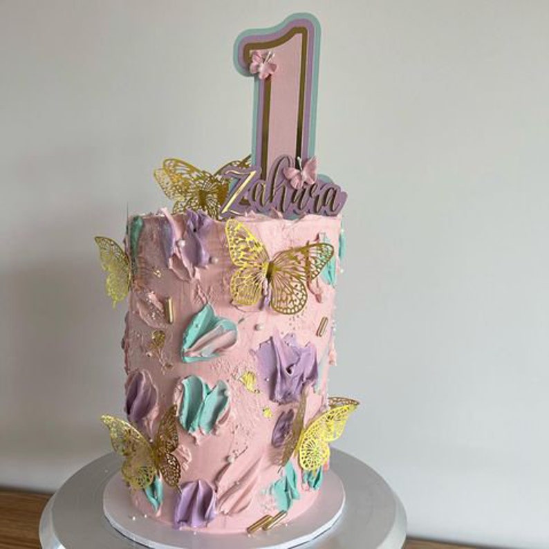 May include: A pink cake decorated with gold butterflies and a purple and green swirl design. The cake is topped with a pink number one and the name "Zahira" in purple.