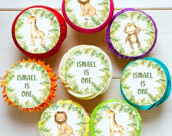 May include: A set of 8 cupcake toppers featuring a jungle theme. The toppers are round and feature various jungle animals, including a lion, giraffe, and monkey. The toppers also include the text "Ismael is one".