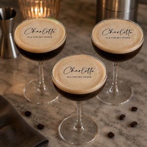 May include: Three clear glass coupe glasses filled with a dark espresso martini cocktail, each topped with a light brown foam layer featuring the printed black text Charlotte IS A TINI BIT OLDER centred on the surface of the drink.