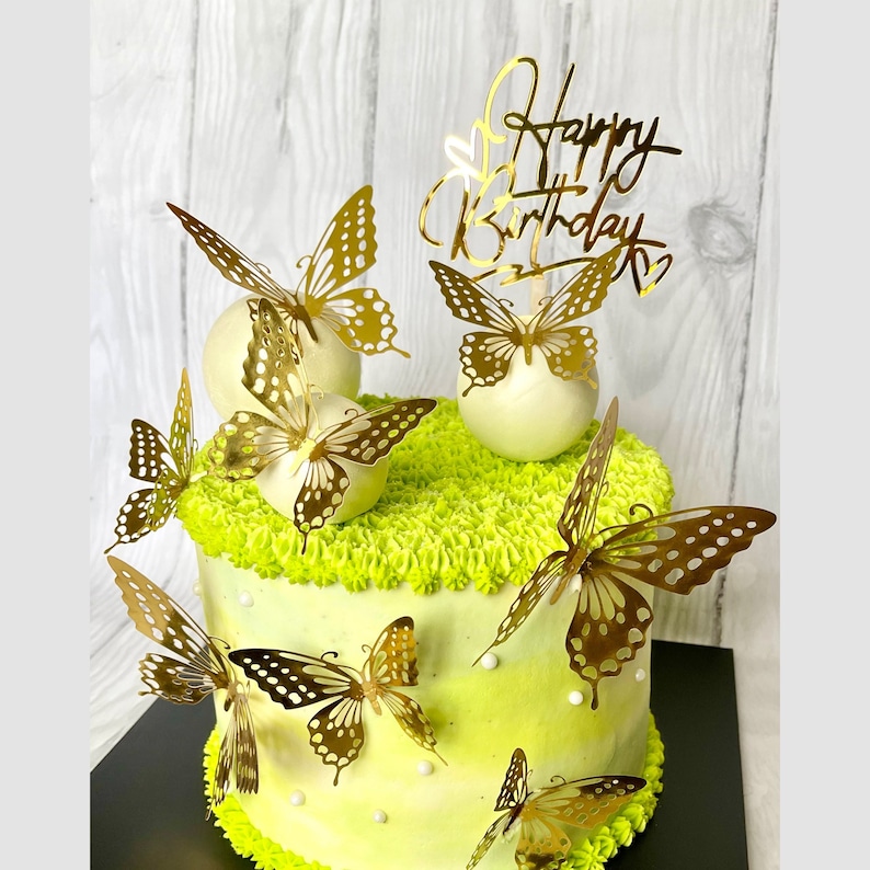 May include: A white cake decorated with green frosting and gold butterfly accents. The cake has a "Happy Birthday" topper and white chocolate balls with gold butterflies on top.