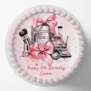 Op de afbeelding: Een roze en witte taart met een aquarelontwerp van make-upkwasten, een cadeaudoos met een roze strik en een make-up palet. De taart is versierd met zilveren glitter en de tekst "Happy 16th Birthday Leonie".