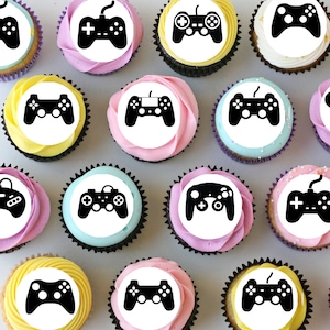 May include: A dozen cupcakes decorated with black and white images of video game controllers. The cupcakes are arranged in a circle on a white surface.