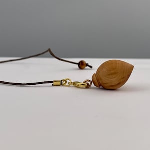Könnte beinhalten: Ein hellbrauner Holzanhänger in Form eines Tropfens oder einer kleinen Muschel, befestigt an einer dunkelbraunen Kordel mit einem goldfarbenen Karabinerverschluss und Biegering. Das Holz zeigt eine natürliche Maserung und eine glatte Oberfläche.