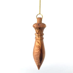 May include: A wooden pendulum with a natural light and dark brown grain pattern, featuring a tapered, pointed shape with a rounded top section and two horizontal grooves, suspended from a thin gold-coloured metal chain by a small circular ring.