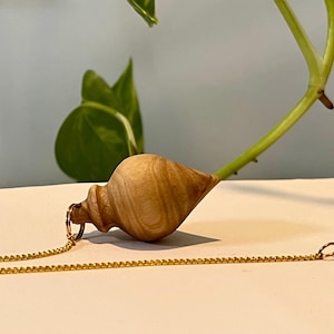 Op de afbeelding: Een houten pendel met een gouden ketting en een kleine, ronde houten kraal aan het uiteinde. De pendel is lichtbruin met een spiraalvormig ontwerp. De achtergrond omvat een groene plant en een wit oppervlak.
