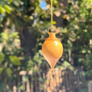 May include: A wooden pendulum with a teardrop shape, suspended by a gold chain. The pendulum is light brown with a pointed tip and a rounded top. The background is a blurred view of green foliage.