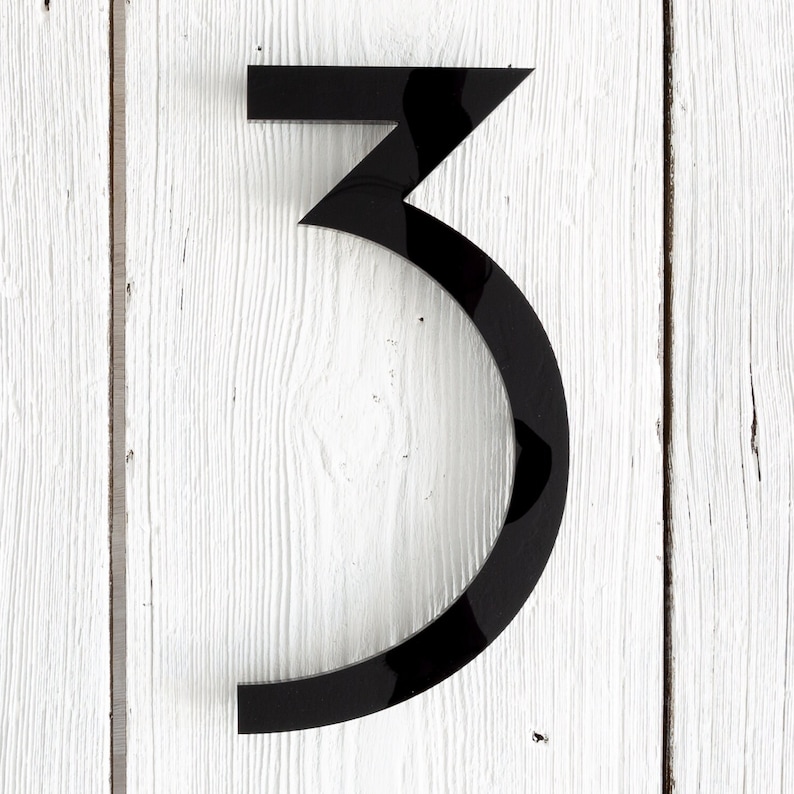 May include: Black number three house number plaque. The number is made of a glossy, black material and is mounted on a white wooden surface.