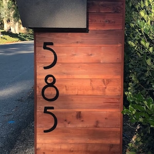 K&ouml;nnte beinhalten: Schwarze Hausnummern aus Metall 585, angebracht an einer Holzwand mit einer rotbraunen Lasur.