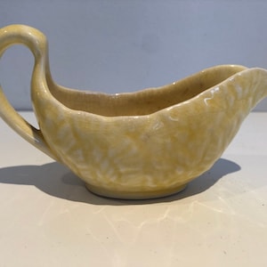May include: A pale yellow ceramic gravy boat with a textured surface and a curved handle.