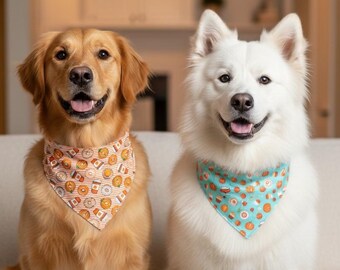 May include: A golden retriever with a tan and white striped bandana featuring a coffee and donut print, and a white fluffy dog with a light blue bandana featuring a pumpkin and autumn treat print, both sitting side by side on a neutral sofa.