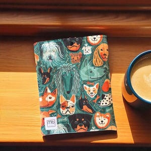 May include: A rectangular fabric book cover featuring a repeating pattern of various dog breeds in shades of orange, black, white, and teal on a dark green background. A small white square label with the black text mb is attached to the bottom left.