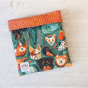 May include: A quilted fabric pouch featuring a colorful pattern of various dog breeds on a teal background. The top flap is orange with small dark dots and a single orange snap button. A small white square label with the text mb is attached.