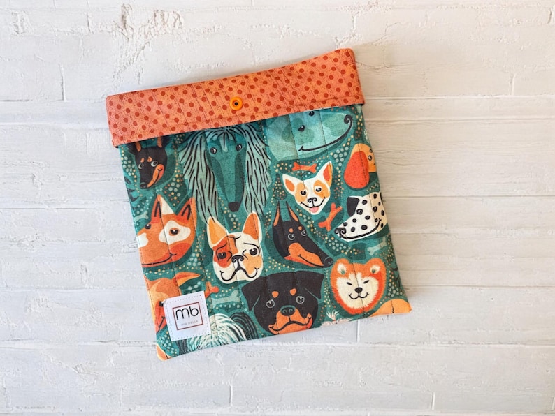 May include: A quilted fabric pouch featuring a colorful pattern of various dog breeds on a teal background. The top flap is orange with small dark dots and a single orange snap button. A small white square label with the text mb is attached.