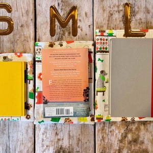 May include: Three fabric book sleeves in small, medium, and large sizes, each featuring a white background with colorful illustrations of dogs, paw prints, and shop fronts. The sleeves hold a yellow book, a paperback book, and a gray book.