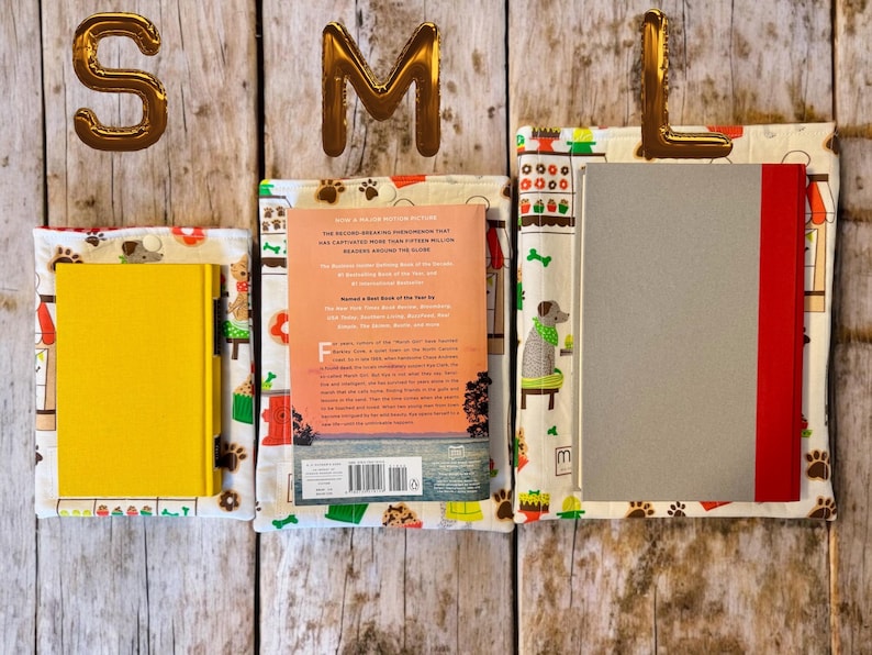 May include: Three fabric book sleeves in small, medium, and large sizes, each featuring a white background with colorful illustrations of dogs, paw prints, and shop fronts. The sleeves hold a yellow book, a paperback book, and a gray book.