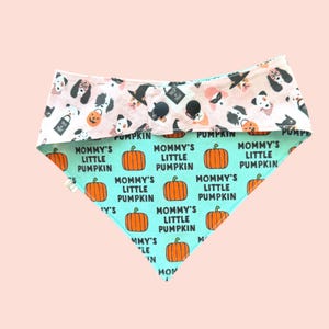 May include: A triangular pet bandana featuring a light pink top section with a pattern of dogs in Halloween costumes and a light green bottom section printed with orange pumpkins and the repeating black text Mommy's Little Pumpkin.
