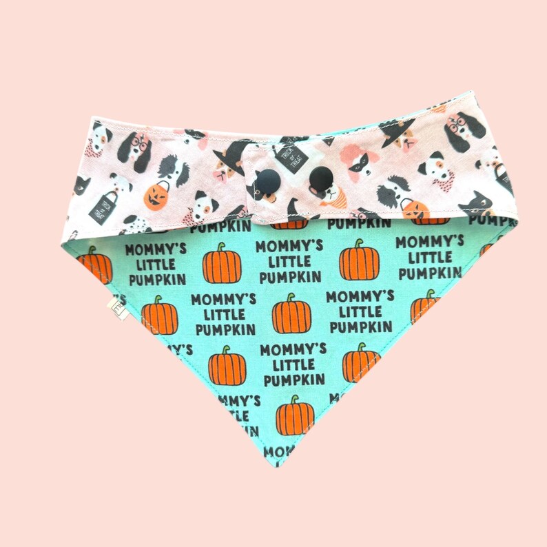 May include: A triangular pet bandana featuring a light pink top section with a pattern of dogs in Halloween costumes and a light green bottom section printed with orange pumpkins and the repeating black text Mommy's Little Pumpkin.