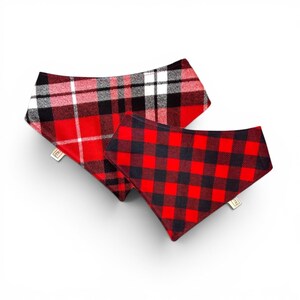 May include: Two dog bandanas in red and black plaid patterns. One bandana has a large plaid design with white accents, while the other features a smaller, square plaid. Both have a tag attached.