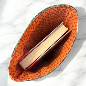 May include: A fabric book sleeve with a green and dark patterned exterior and an orange interior lining featuring a small brown polka dot print. The sleeve is open and contains a single hardcover book with a red spine and cream-colored pages.