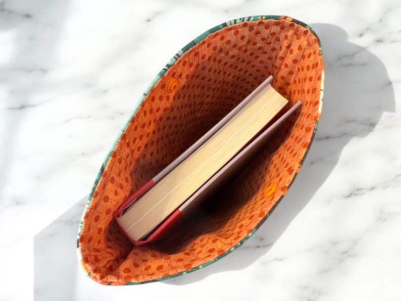 May include: A fabric book sleeve with a green and dark patterned exterior and an orange interior lining featuring a small brown polka dot print. The sleeve is open and contains a single hardcover book with a red spine and cream-colored pages.