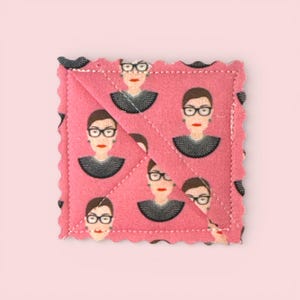 May include: Pink square coaster with a repeating pattern of a woman with glasses, a black collar, and red lipstick. The coaster has a scalloped edge and a quilted design.