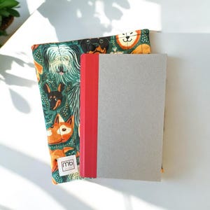 May include: A fabric book sleeve featuring a repeating pattern of various dog breeds on a dark green background, with a small white label reading mb. A gray notebook with a red spine is placed on top of the quilted sleeve, which has a button closure.