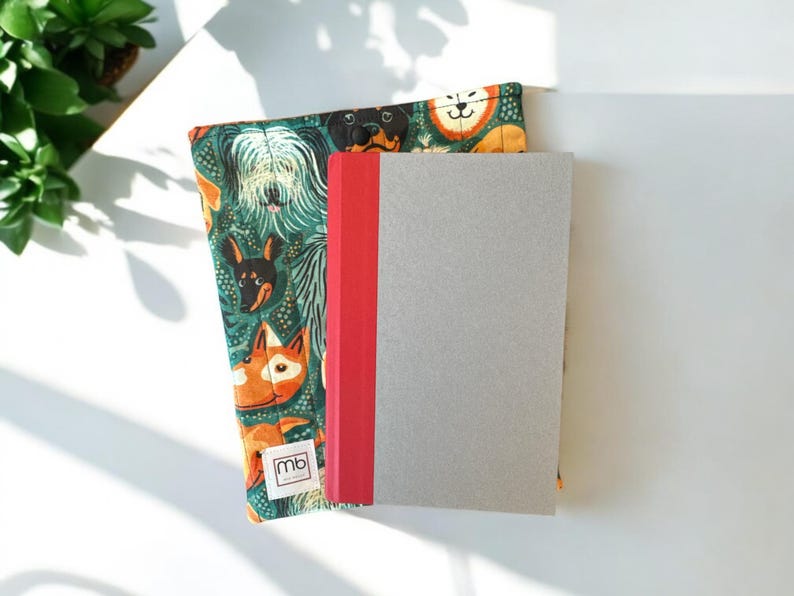 May include: A fabric book sleeve featuring a repeating pattern of various dog breeds on a dark green background, with a small white label reading mb. A gray notebook with a red spine is placed on top of the quilted sleeve, which has a button closure.