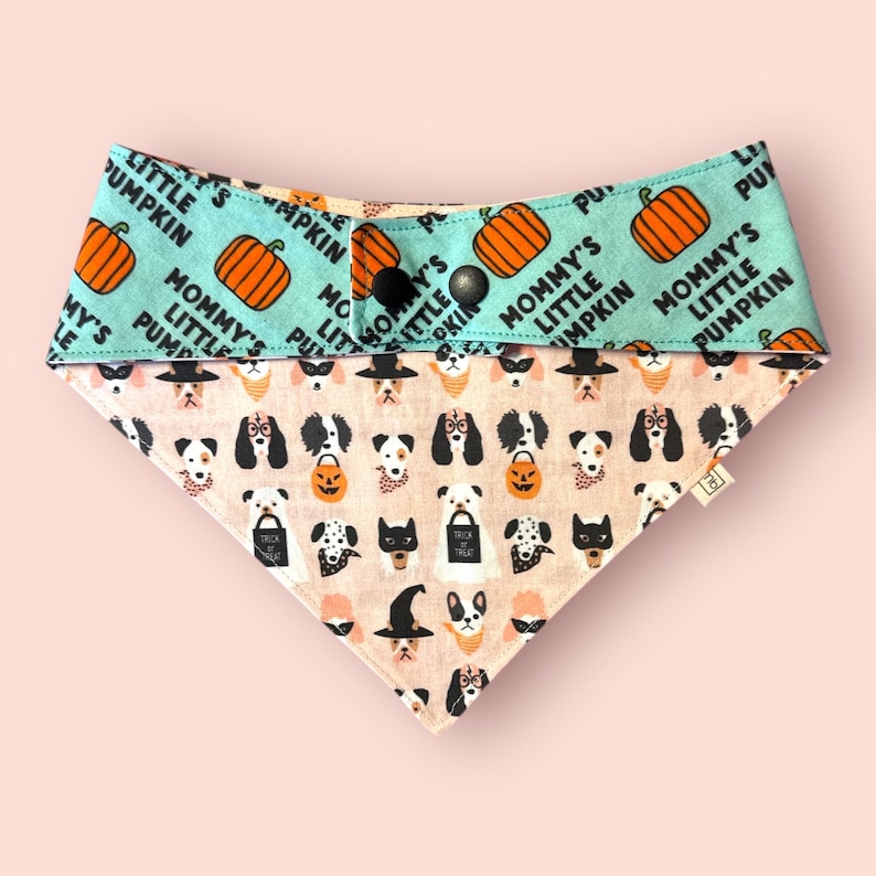 May include: A triangular pet bandana featuring a light blue top section with orange pumpkin illustrations and the text Mommy's Little Pumpkin. The bottom section is beige with a repeating pattern of dogs in Halloween costumes and trick-or-treat bags.