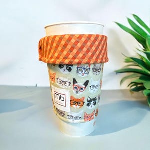 May include: A reusable coffee cup cozy with a plaid orange and white top and a bottom featuring a pattern of cats wearing glasses. The text "mb" is visible on the cozy.