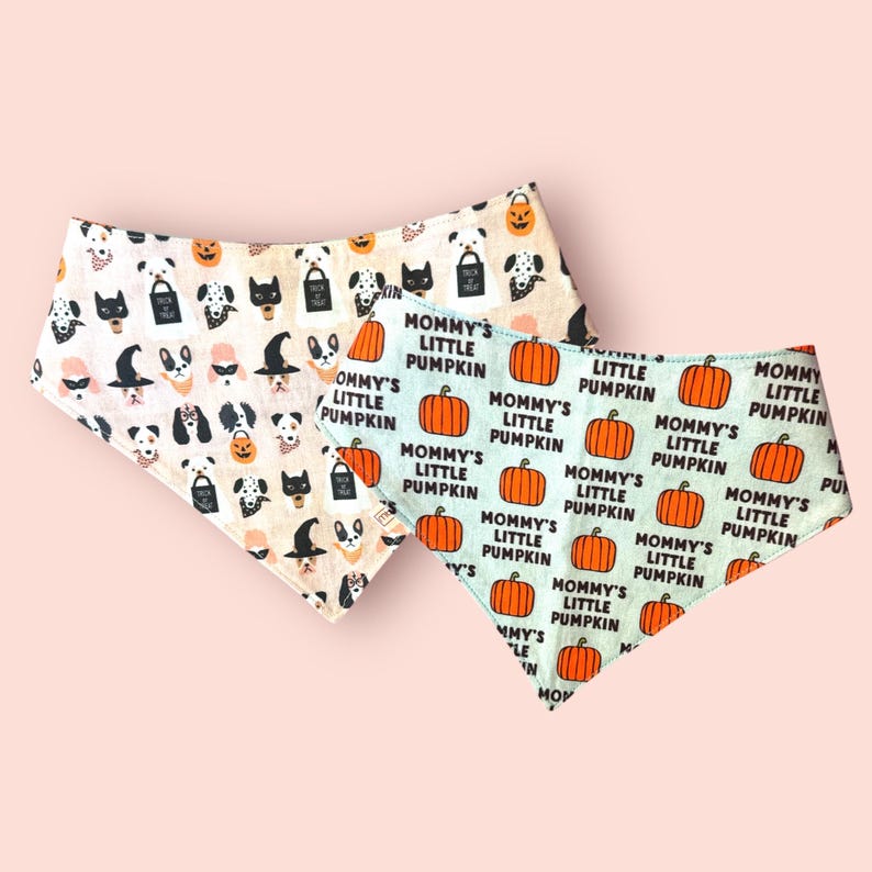 May include: Two triangular pet bandanas. One features a cream background with a repeating pattern of dogs in Halloween costumes and trick-or-treat bags. The other is light blue with a repeating pattern of orange pumpkins and the text Mommy's Little Pumpkin.