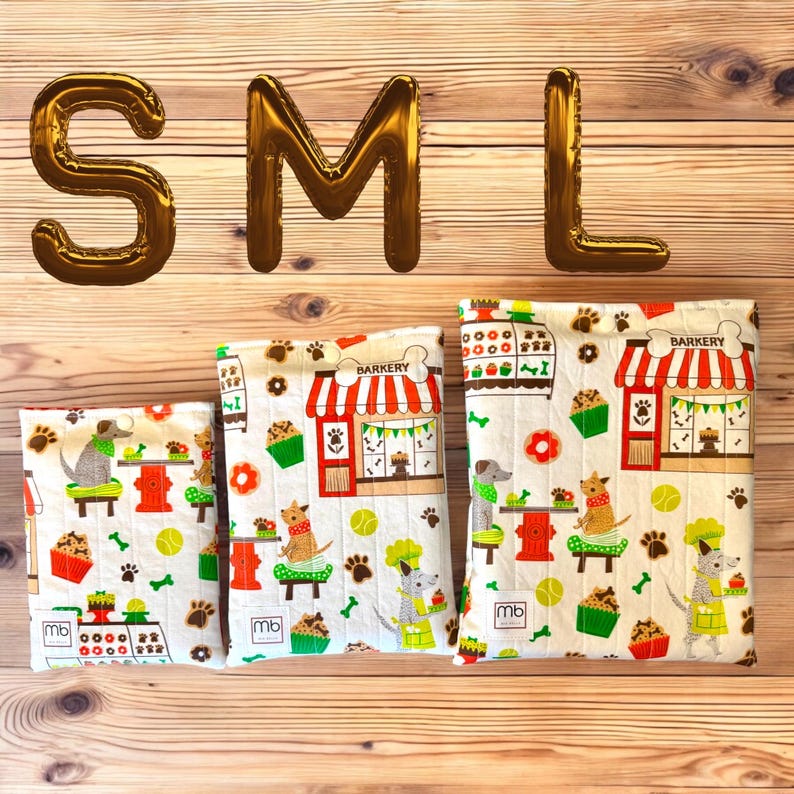 May include: Three rectangular fabric pouches in small, medium, and large sizes, each featuring a white background with a colorful pattern of dogs, cupcakes, and a bakery storefront labeled BARKERY. Each pouch has a small white label with mb.