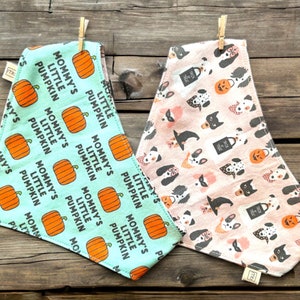 May include: Two triangular pet bandanas. The left one is light blue with orange pumpkins and the text "MOMMY'S LITTLE PUMPKIN" in black. The right one is light pink with a pattern of various dog breeds, witch hats, and trick-or-treat bags.