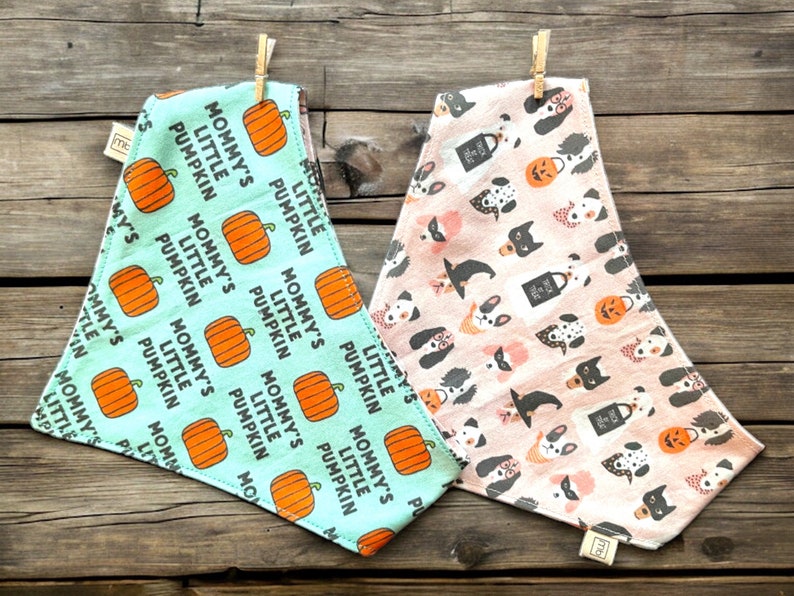 May include: Two triangular pet bandanas. The left one is light blue with orange pumpkins and the text "MOMMY'S LITTLE PUMPKIN" in black. The right one is light pink with a pattern of various dog breeds, witch hats, and trick-or-treat bags.