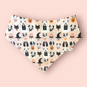 May include: A triangular pet bandana in a light beige fabric featuring a repeating Halloween pattern of dogs wearing costumes, witch hats, and masks, alongside orange jack-o'-lanterns and black trick-or-treat bags with the text Trick or Treat.