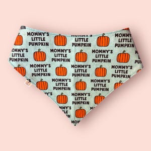 May include: A triangular bandana with a light blue background featuring a repeating pattern of orange pumpkins and the black text Mommy's Little Pumpkin. The fabric has a small white tag on the left side and a pointed bottom edge.